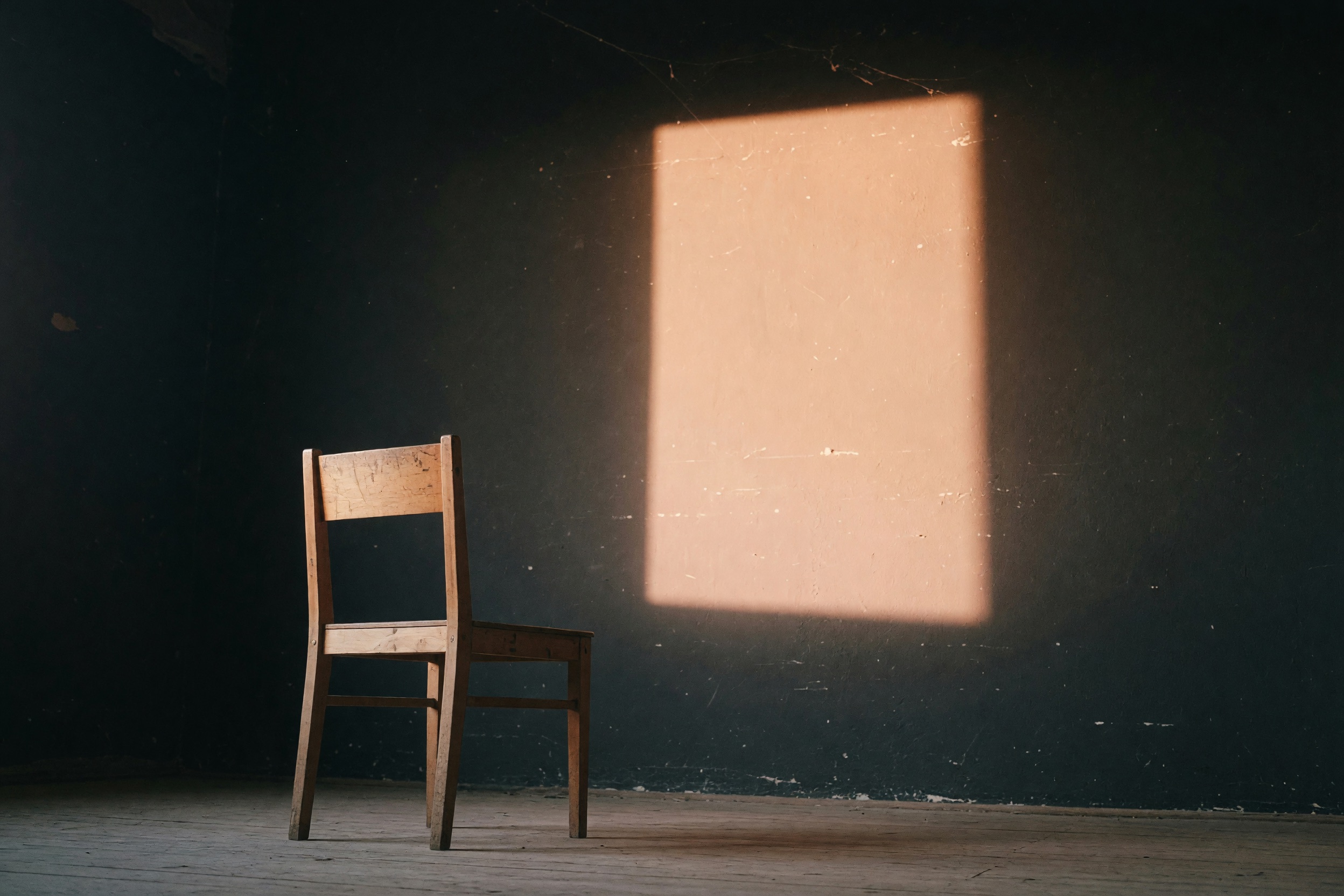 An empty chair facing a warm rectangle of light on a dark wall